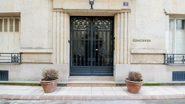 A building entrance with a black ornate door, two potted plants, and the name "CONCIERGE" on the wall. - Appartement Carpette - Welkeys (Paris)