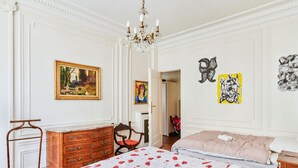 A bedroom with a large bed, a wooden dresser, a chair, and a chandelier. - Appartement Carpette - Welkeys (Paris)