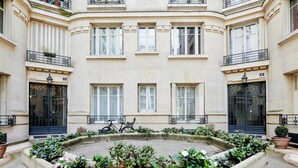 A courtyard with a central fountain surrounded by flower beds, featuring a building with multiple windows and balconies. - Appartement Carpette - Welkeys (Paris)