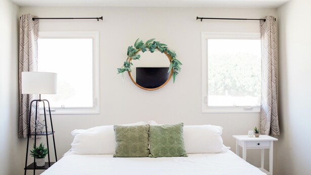 A bedroom with a bed, two pillows, a bedside table with a lamp, a small plant, and a wall-mounted mirror with a wreath.