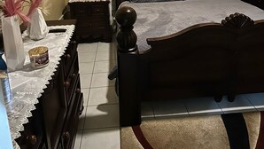 Et soveværelse med en træseng, et natbord med lampe, en kommode og et vindue med blomstrede gardiner.