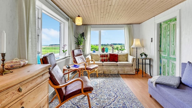 Ein gemütliches Wohnzimmer mit Holzbalkendecke, großen Fenstern mit Blick ins Grüne, einer Couch, einem Sessel und einem kleinen Tisch.