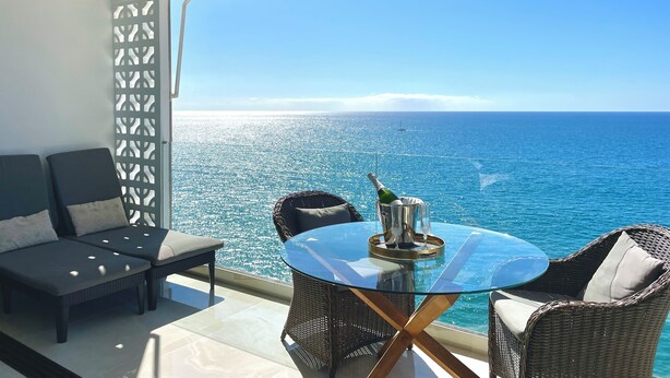 A balcony with a glass table, wicker chairs, and a sofa overlooking a clear blue sea. - Seaside Bliss: Spectacular Views, Beachfront Haven (Patalavaca)
