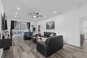 Uma sala de estar moderna com um sofá de couro preto, uma mesa de centro e uma TV. Há uma varanda com vista para o exterior.