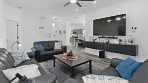 A modern living room with a ceiling fan, a flat-screen TV, a dark wooden coffee table, and a black sofa with patterned cushions. - 4 Bedrooms/2 Bathrooms AMAZING HOME! PRIVATE POOL! (Kissimmee)