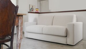Un canapé blanc dans une pièce où se trouvent une chaise en bois et une petite table sur laquelle sont posés des livres et un tableau.