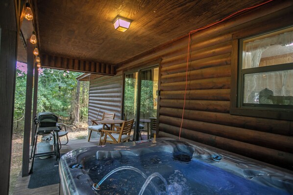 A wooden cabin with a hot tub, a table with chairs, and a window overlooking greenery.