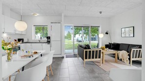 Eine moderne Küche mit weißen Schränken, ein Essbereich mit weißem Tisch und Stühlen, eine schwarze Couch und ein großes Fenster mit Blick ins Freie.