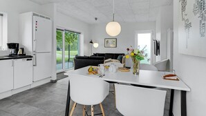 Eine moderne Küche mit weißen Schränken, ein Essbereich mit weißem Tisch und Stühlen sowie ein großes Fenster mit Blick ins Freie.