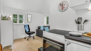 Une cuisine moderne avec des meubles blancs, un plan de travail noir et un four encastré. On y trouve une fenêtre donnant sur la verdure, une chaise avec une serviette bleue et une horloge au mur.