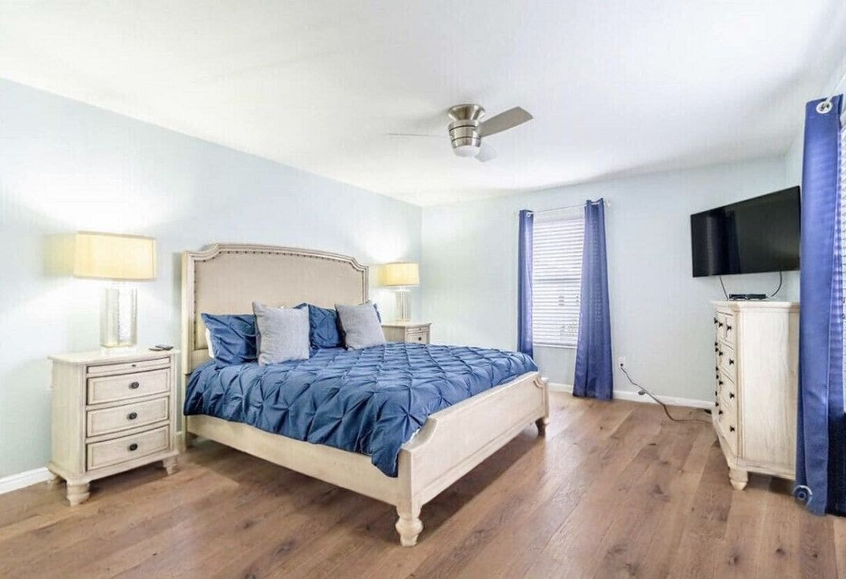 Une chambre à coucher comprenant un lit, des tables de chevet, un ventilateur de plafond, une télévision et des rideaux.