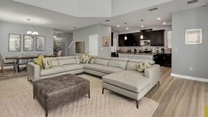 A spacious living room with a large sectional sofa, a dark ottoman, and a dining area with a glass table. A kitchen with dark cabinets and a central island is visible in the background.