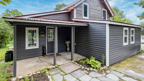A two-story house with a dark exterior, a red roof, and a front porch with a welcome sign. - Cozy Adirondack Loft Near Lake Champlain & George, Hiking and Historical Sites (Ticonderoga)