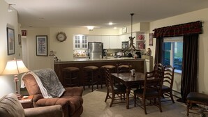 A cozy living room with a brown sofa, a wooden dining table with chairs, a kitchen with white cabinets and stainless steel appliances, and a window with curtains. - Winterplace Condo Steps away from slopes (Ludlow)