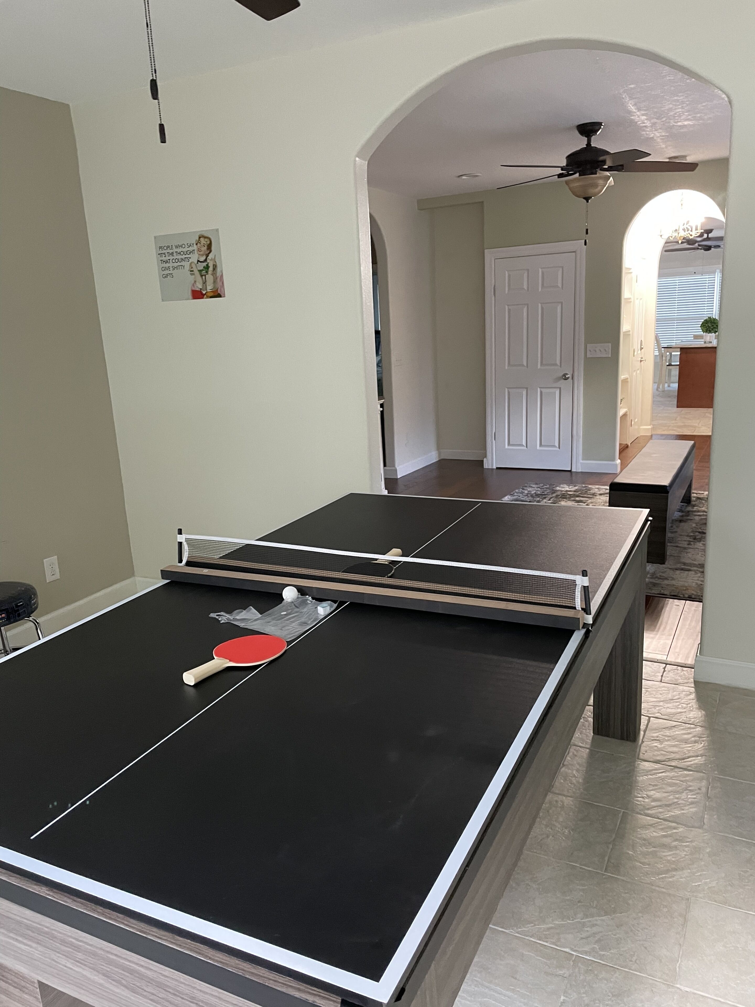 Une table de ping-pong se trouve dans une pièce avec une porte blanche, un ventilateur de plafond et un couloir menant à une cuisine.