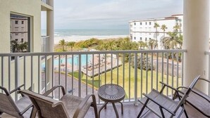 A balcony with chairs and a table overlooking a beach and a pool area.
