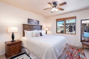 Une chambre à coucher avec un lit, deux lampes de chevet, une table de chevet en bois, un ventilateur de plafond, une fenêtre avec vue et un tapis à motifs.