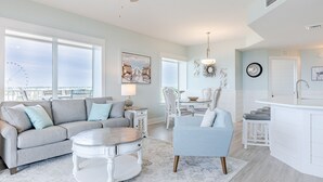 A spacious living room with a gray sofa, a round coffee table, and a blue armchair. A dining area with a white table and chairs is visible in the background. - End-Unit 3Bed/3Bath Condo w/ Gulf Views, Wraparound Balcony & Tiki Bar! (Panama City Beach)