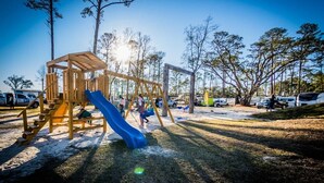 A playground with a slide and climbing structure, surrounded by trees and parked cars. - Biloxi Bay Adventure: Stay in Our Comfortable Camper Rental (Biloxi)