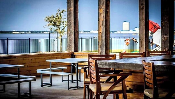 An outdoor dining area with wooden tables and chairs, a view of the water, and a distant building. - Biloxi Bay Adventure: Stay in Our Comfortable Camper Rental (Biloxi)
