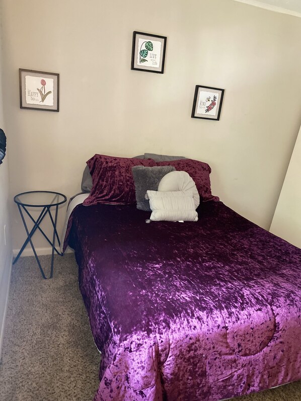A bedroom with a purple bedspread, a small round table, and three framed pictures on the wall.