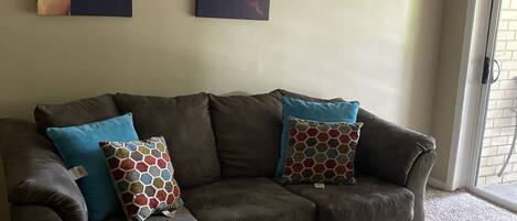 A living room with a brown sofa, patterned cushions, a glass coffee table, and abstract artwork on the wall.