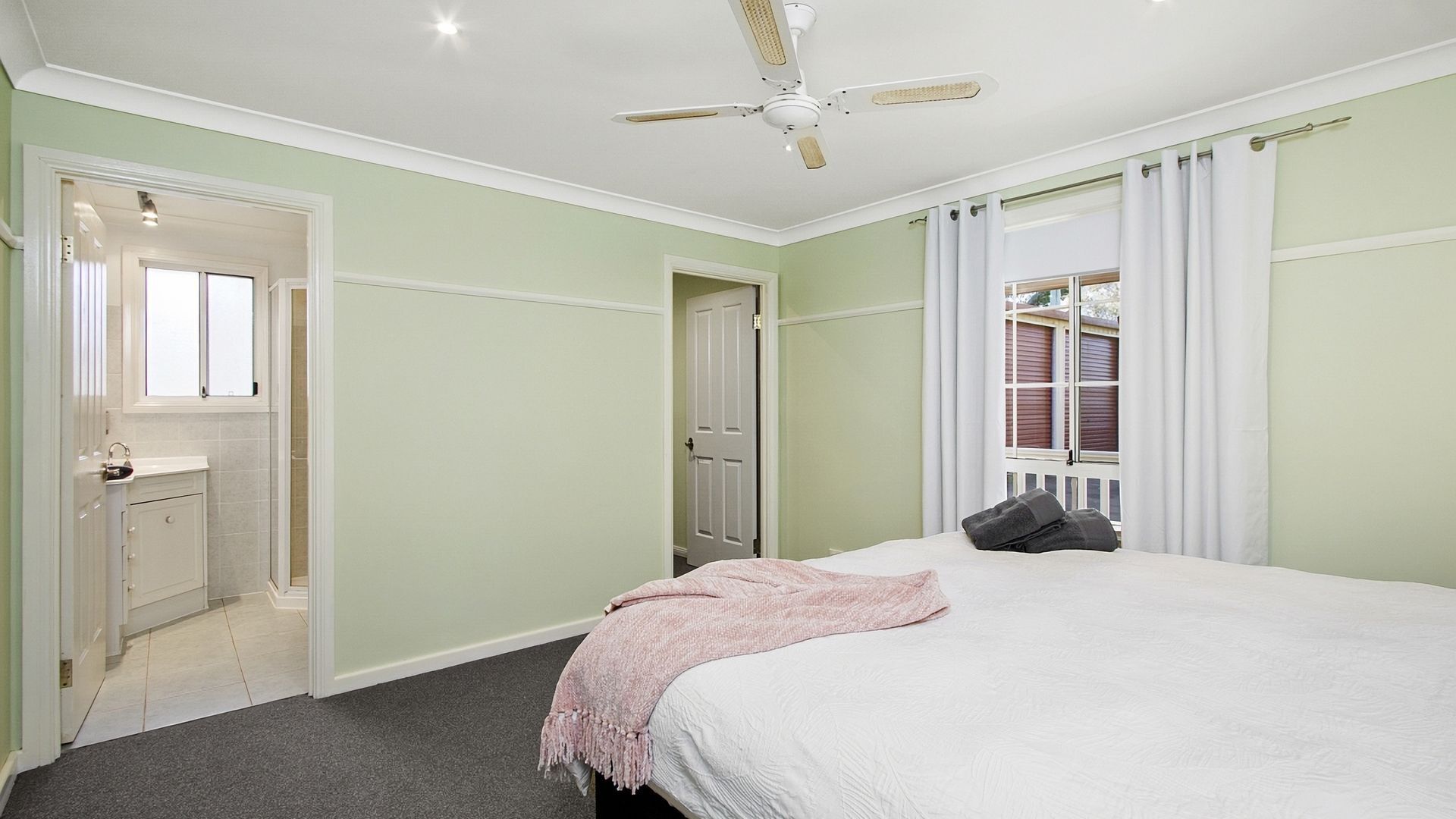 Une chambre à coucher comprenant un lit, un ventilateur de plafond, une fenêtre avec des rideaux et une porte donnant sur une salle de bain.