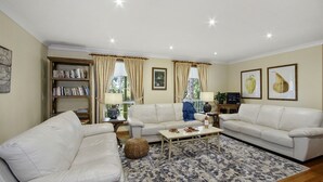 A living room with two white sofas, a coffee table, a bookshelf, and a large window with curtains. - HILLDO - Beautiful home set amongst the tree  - SPECIALS ON NOW ! (Kurrajong Heights)