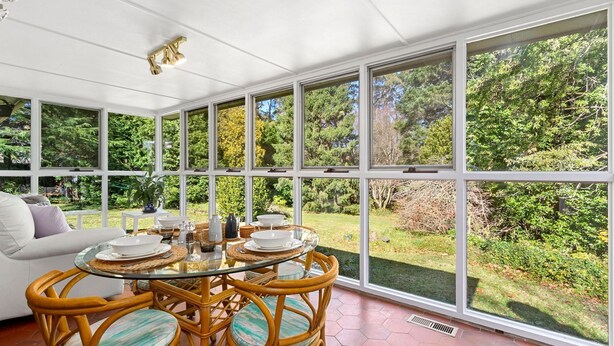 A conservatory with a glass table set for dining, surrounded by wicker chairs, and a white sofa.