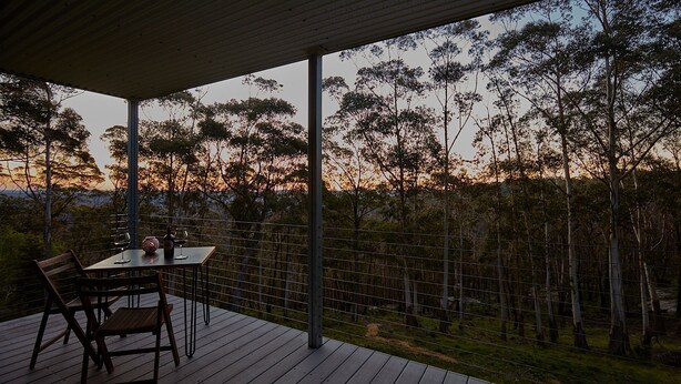 Área de refeições ao ar livre com mesa e cadeiras, cercada por um deck de madeira e uma cerca de arame, com vista para uma paisagem arborizada.