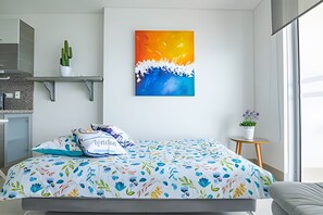 Een moderne slaapkamer met een bed, een kleurrijk bloemenbedekking, een schilderij aan de muur, een cactus op een plank en een klein tafeltje met een plant.