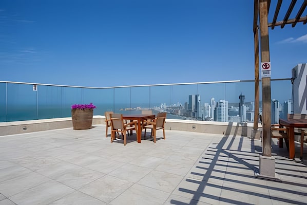 Een terras op het dak met een tafel, stoelen, een houten plantenbak en uitzicht op de skyline van de stad.