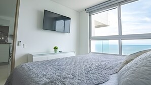 Ein modernes Schlafzimmer mit einem großen Bett, einem an der Wand befestigten Fernseher, einer kleinen Pflanze auf einem weißen Konsolentisch und Meerblick durch ein großes Fenster.