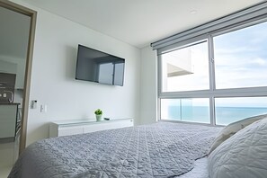 Quarto moderno com uma cama grande, televisão montada, uma pequena planta sobre um console branco e vista para o mar através de uma grande janela.
