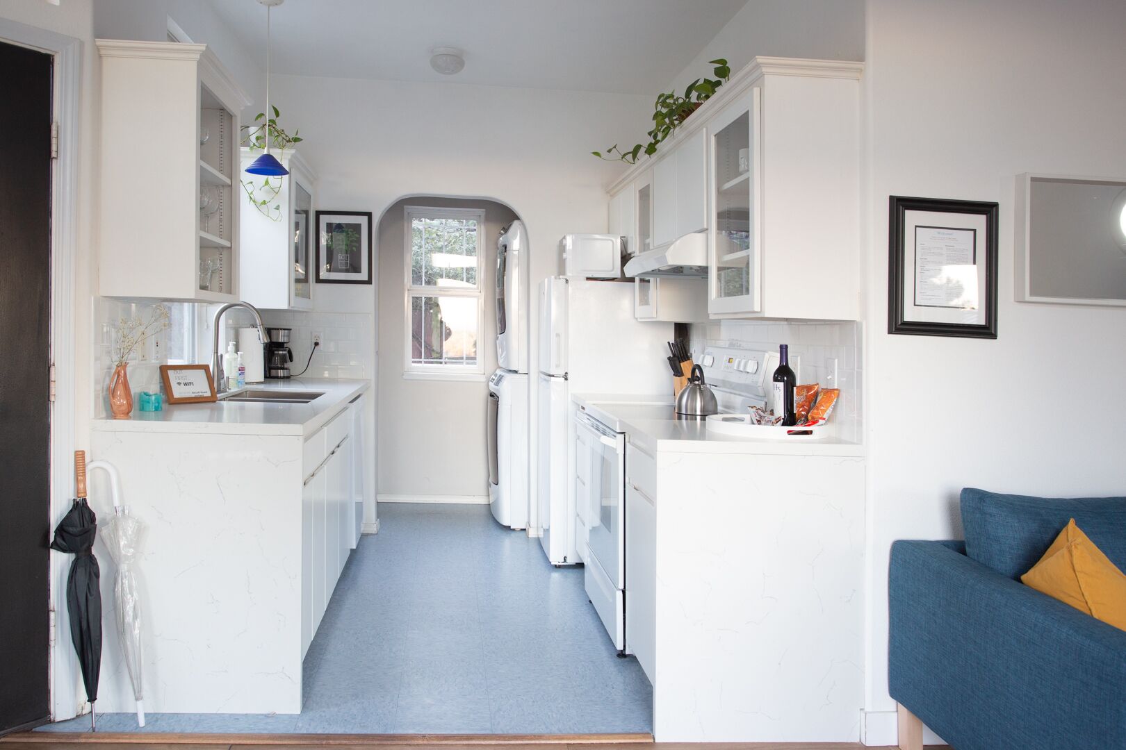 A narrow kitchen with white cabinets, a blue countertop, and a blue sofa in the adjacent living area.
