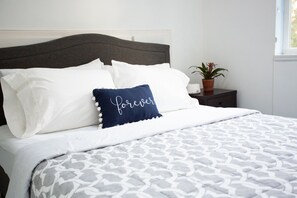 Un lit bien fait, avec une literie blanche et grise à motifs, une tête de lit gris foncé et un coussin décoratif orné du mot « toujours ».