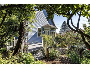 Une maison à l’extérieur bleu, dotée d’un porche abrité et d’un jardin où poussent diverses plantes et fleurs.