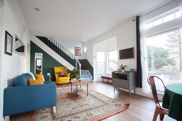 Un salon spacieux avec un canapé bleu, un fauteuil jaune et une grande fenêtre donnant sur l’extérieur.