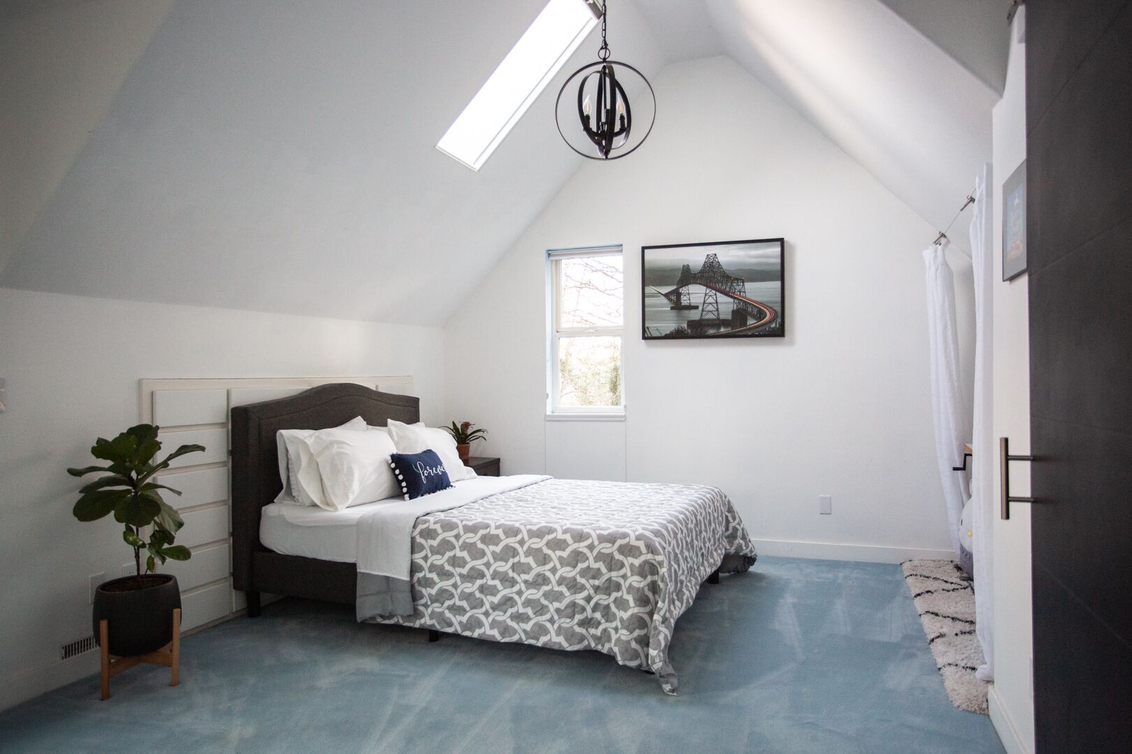 A bedroom with a sloped ceiling, a bed with patterned bedding, a potted plant, a framed picture on the wall, and a window.