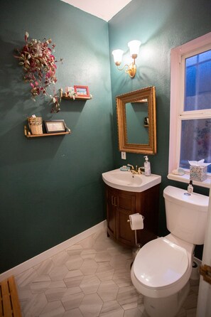 Une salle de bain avec un mur vert, un lavabo blanc, un meuble en bois, des toilettes et une fenêtre donnant sur un beau paysage.