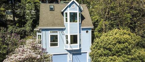 A two-story house with a gabled roof, a front garage, and surrounded by dense trees and shrubs.