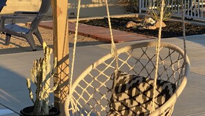 Un fauteuil suspendu en forme de hamac, avec un cactus à côté, un banc et une zone pavée, le tout agrémenté d’une clôture et de plantes en arrière-plan.