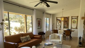 Un salon avec un canapé en cuir marron, un fauteuil blanc et une table basse en verre. De grandes fenêtres sont ornées de rideaux, un ventilateur de plafond est installé et des œuvres d’art décorent les murs.