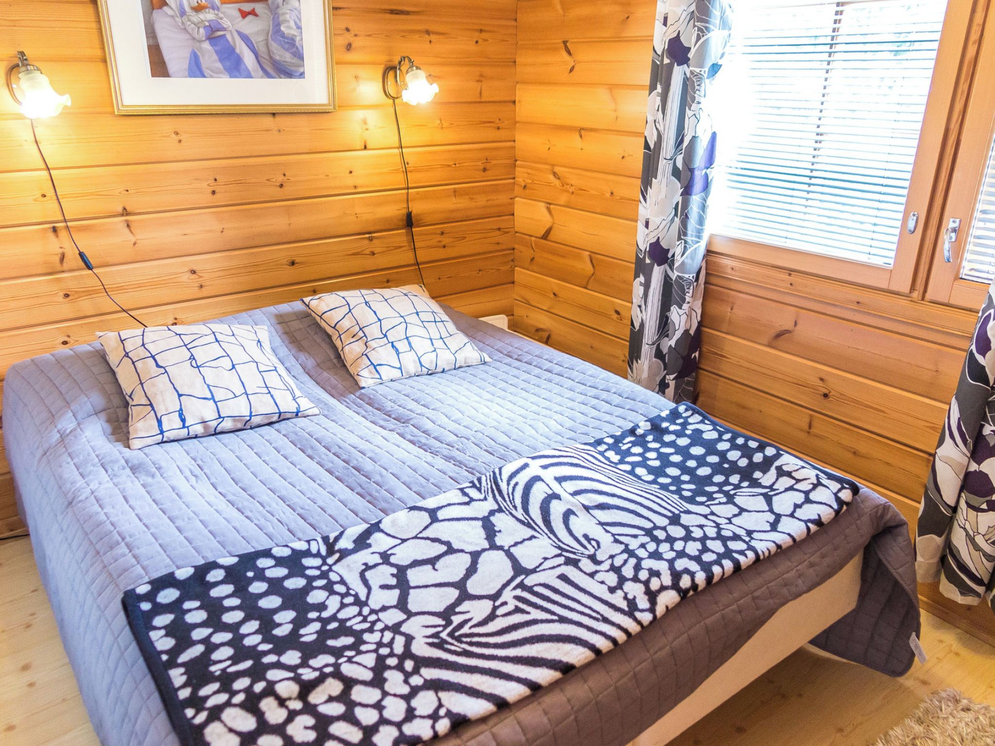 Habitación de cabaña de madera con cama, dos almohadas, un cuadro en la pared y una ventana con persianas.