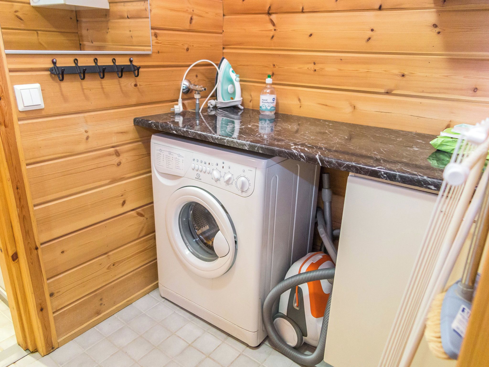Un espace réduit dédié à la buanderie, comprenant un lave-linge, un fer à repasser et un flacon de gel hydroalcoolique.