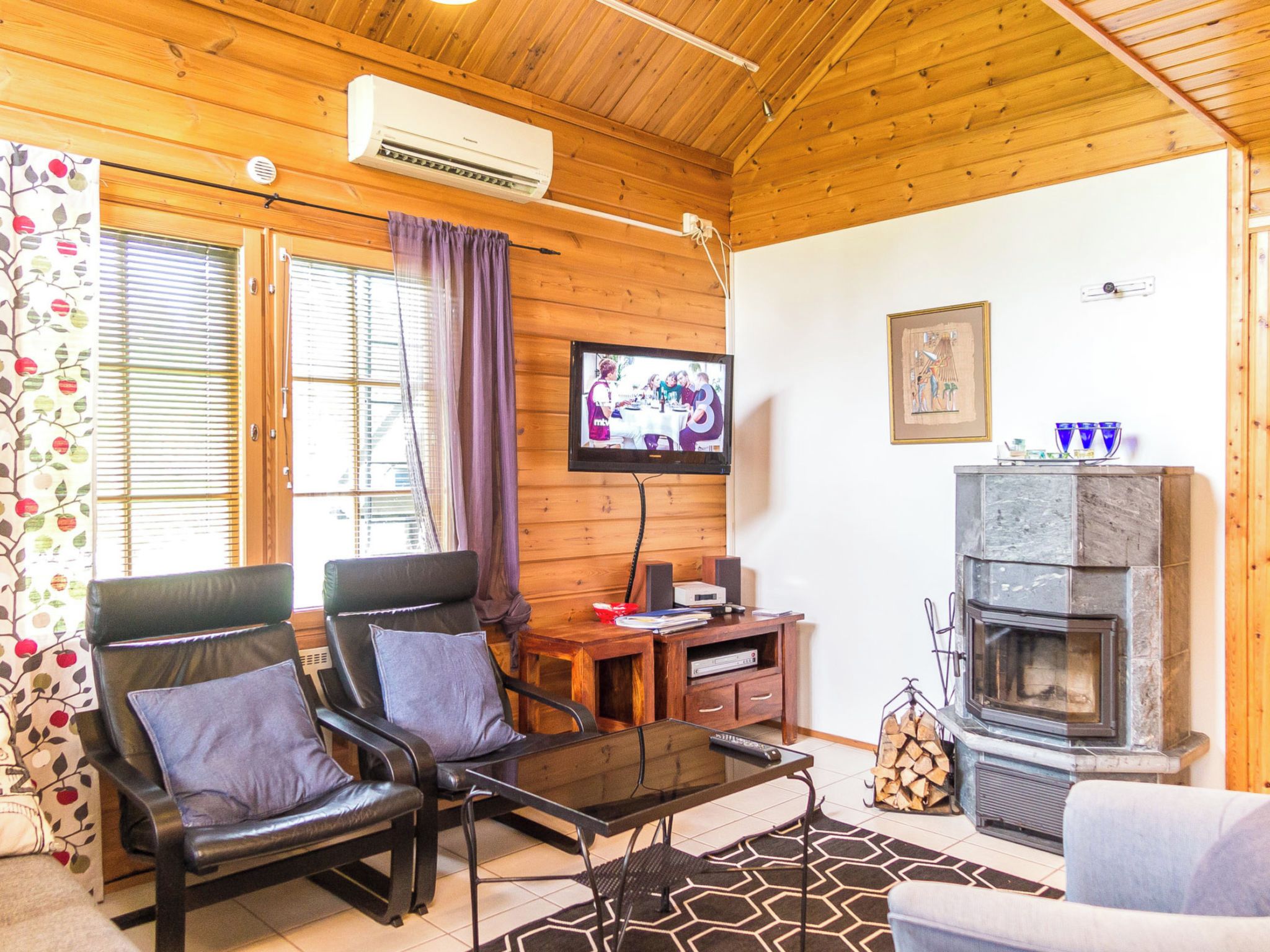 A cozy living room with a fireplace, leather chairs, a flat-screen TV, and a wooden ceiling.