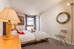 A bedroom with a bed, a wooden desk with a lamp, a chair, a round mirror, and a window with curtains.