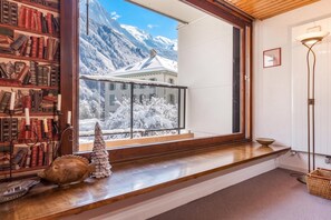 A room with a large window overlooking a snowy mountain landscape, a bookshelf filled with books, and a small Christmas tree.