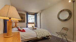 Une chambre à coucher comprenant un lit, une table de chevet en bois, une lampe, une chaise, une fenêtre avec des rideaux, un miroir fixé au mur et un radiateur.