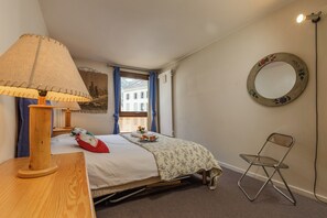 Ein Schlafzimmer mit einem Bett, einem Nachttisch aus Holz, einer Lampe, einem Stuhl, einem Fenster mit Vorhängen, einem wandmontierten Spiegel und einem Heizkörper.